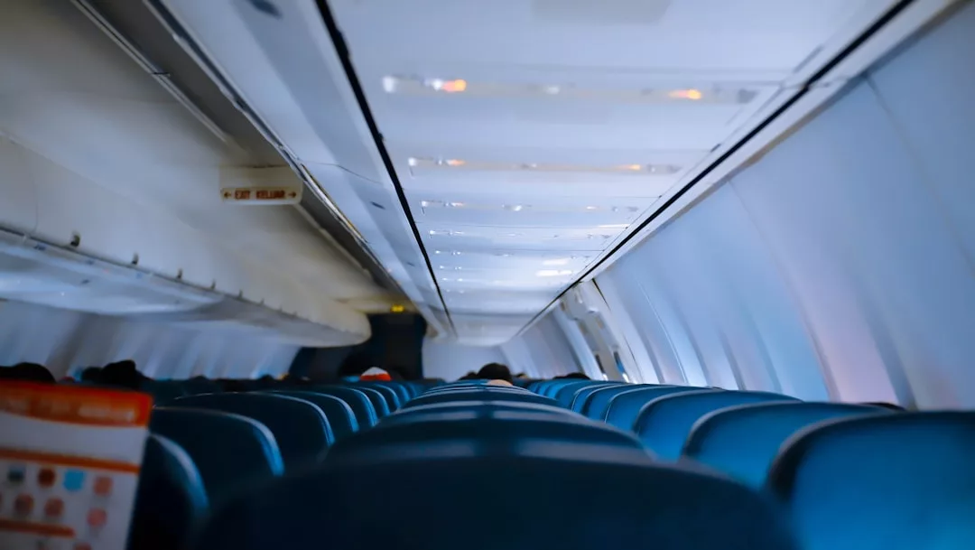 Das Innere eines Flugzeugs zeigt Reihen von blauen Passagiersitzen und Gepäckfächern, wobei einige Lichter eingeschaltet sind und eine Sicherheitskarte im Vordergrund zu sehen ist, die wichtige Sicherheitsmaßnahmen für Alleinreisende hervorhebt.