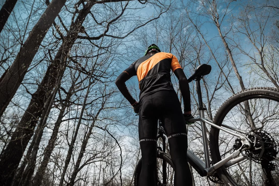 Ein Radfahrer in schwarz-oranger Kleidung steht neben einem Mountainbike und blickt auf hohe, blattlose Bäume vor blauem Himmel - perfekte Bedingungen für sicheres Training und Outdoor-Training in der Natur.