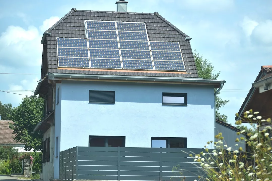 Ein zweistöckiges Haus mit blauen Wänden zeigt den Solarenergie-Trend, mit Sonnenkollektoren auf dem dunklen Ziegeldach, einem Zaun und Nachbarhäusern in Sichtweite.