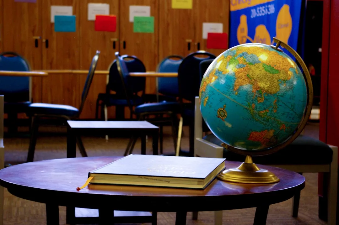 Ein Globus und ein geschlossenes Buch liegen auf einem runden Tisch in einem Klassenzimmer mit Stühlen, einer Pinnwand und bunten Papieren an der Wand - genau das Richtige für diejenigen, die sich auf Sprachkurse im Ausland vorbereiten, um ihre Sprachbeherrschung zu verbessern.