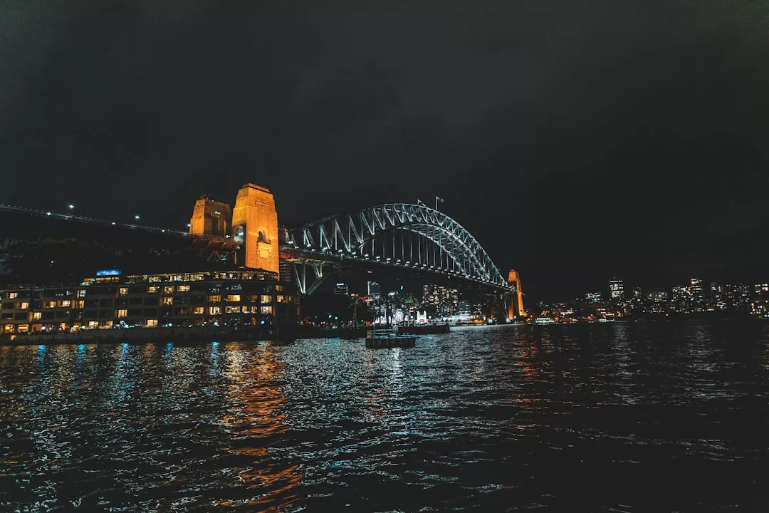 Die Sydney Harbour Bridge bei Nacht, ein urbanes Highlight von Down Under, erstrahlt im Licht der Lichter der Stadt, die sich auf dem Wasser spiegeln, eingerahmt von Gebäuden und dem dunklen Himmel im Hintergrund.