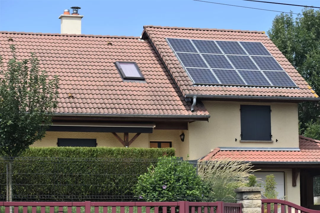 Ein beigefarbenes Haus mit roten Dachziegeln und Sonnenkollektoren auf dem oberen Dach ist ein Beispiel für Energieeffizienz, mit geschlossenen Fensterläden, Grünpflanzen und einem roten Zaun.