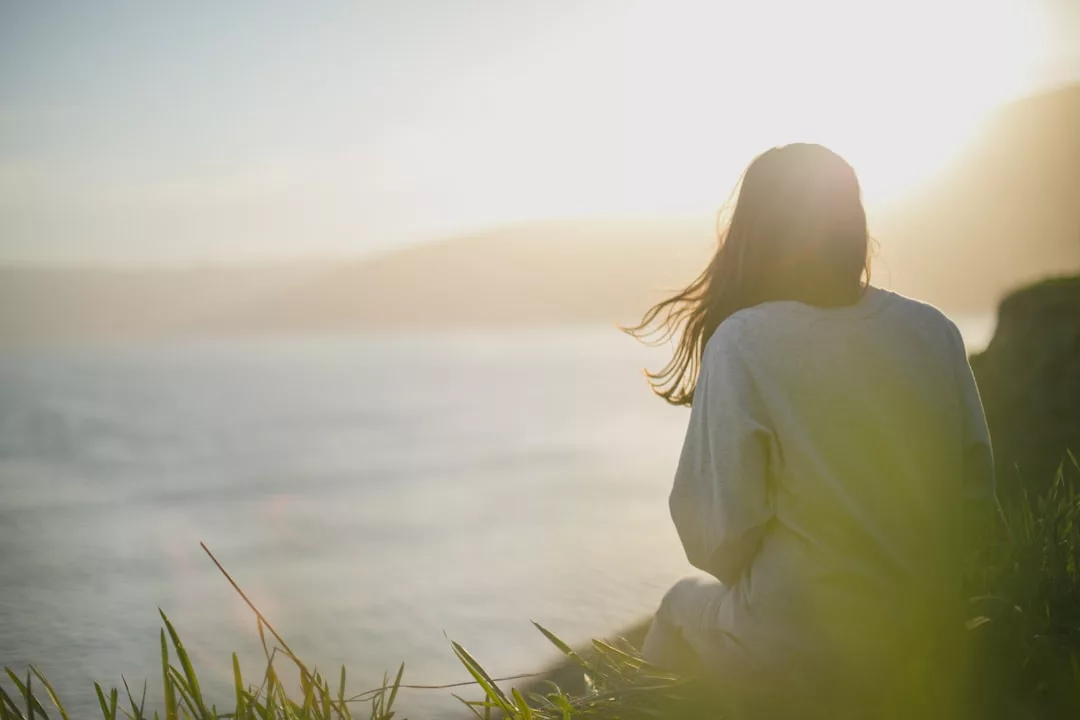 Eine Person, die im Gras in der Nähe des Wassers sitzt, mit Blick auf einen hellen Sonnenaufgang oder Sonnenuntergang mit Bergen in der Ferne - ein Moment der Stressbewältigung und inneren Ruhe.