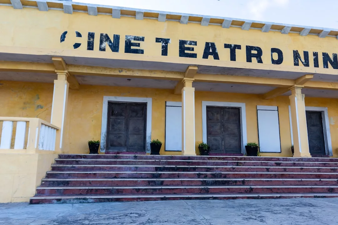 Ein gelbes Gebäude mit verblassten Wänden, drei verschlossenen Holztüren, Topfpflanzen und dem teilweise sichtbaren Schild "CINE TEATRO NIN" über dem Eingang deutet auf einen Ort hin, an dem einst Vielfalt und Bühnenkunst während Theaterfestivals lebendig waren.