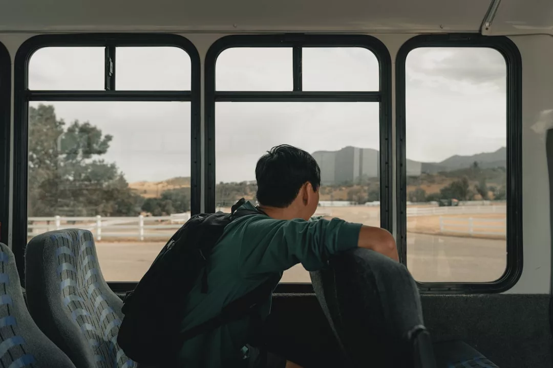 Ein Soloabenteurer mit Rucksack sitzt allein auf einem Bussitz und blickt aus dem Fenster auf eine Landschaft mit Bergen und einem Zaun im Hintergrund. Perfekt für Alleinreisende, die neue Reisetipps suchen.