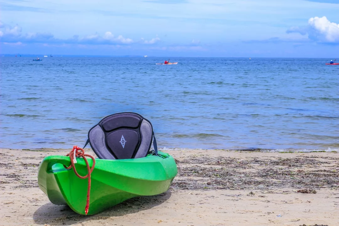 Ein grünes Kajak mit schwarzem Sitz ruht auf dem Sandstrand in der Nähe der Küste, ruhiges blaues Meerwasser und weit entfernte Boote sind unter einem teilweise bewölkten Himmel zu sehen - perfekt für Fotografie Tipps oder jeden, der fotografieren lernen möchte.