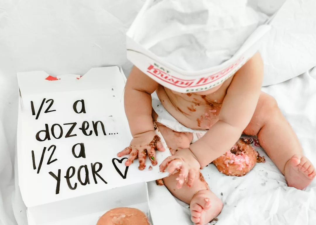 Ein Baby sitzt auf einer weißen Decke mit Donuts und einer geöffneten Krispy-Kreme-Schachtel, auf der "1/2 Dutzend... 1/2 ein Jahr." Um das erste Jahr als Eltern zu feiern, hat das Baby Donut-Glasur an Händen und Körper.