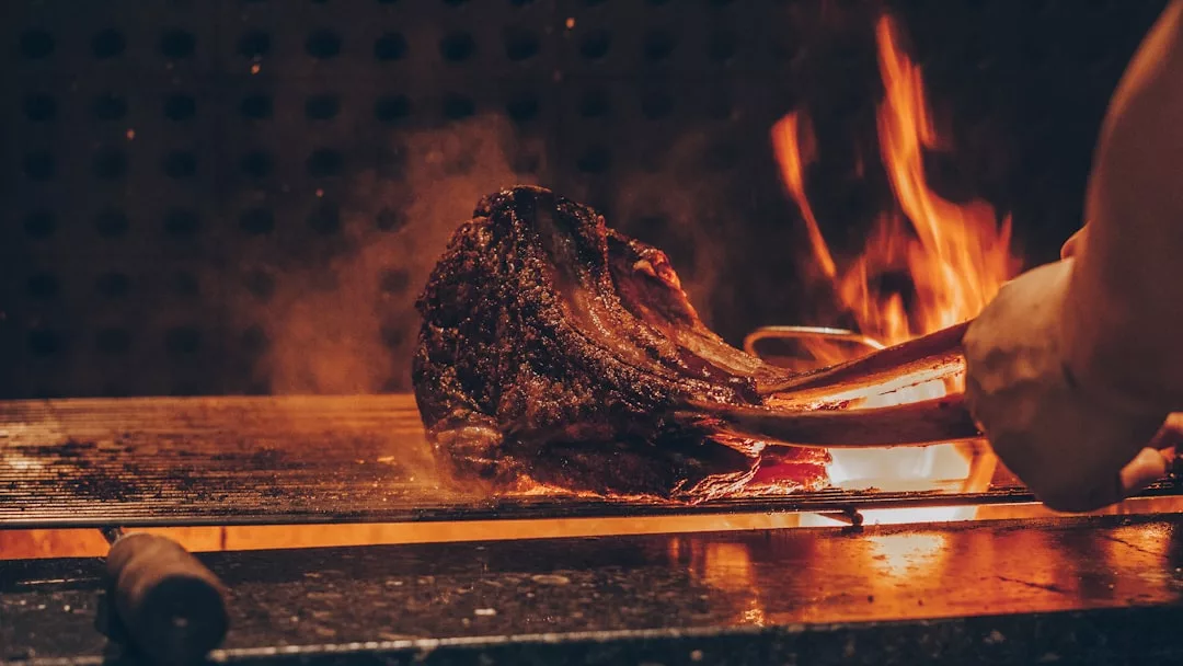Eine Person grillt ein saftiges Steak über einer offenen Flamme mit einer Zange, mit sichtbarem Feuer und Rauch im Hintergrund.