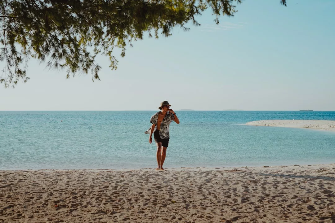 Eine Person auf einer Solo-Reise, die einen Hut und ein gemustertes Hemd trägt, geht an einem Sandstrand in der Nähe des Ozeans spazieren, wobei sie eine Tasche und Schuhe trägt, während über ihr die Äste der Bäume und das ruhige blaue Wasser im Hintergrund zu sehen sind.