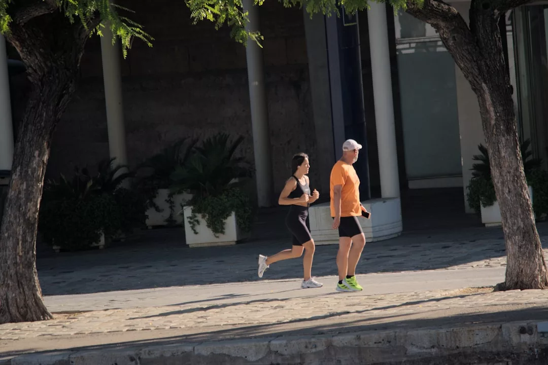 Zwei Personen joggen auf einem sonnenbeschienenen Bürgersteig in der Nähe von Bäumen und großen Topfpflanzen, möglicherweise mitten in ihrem Halbmarathon-Training, mit einem Gebäude im Hintergrund.