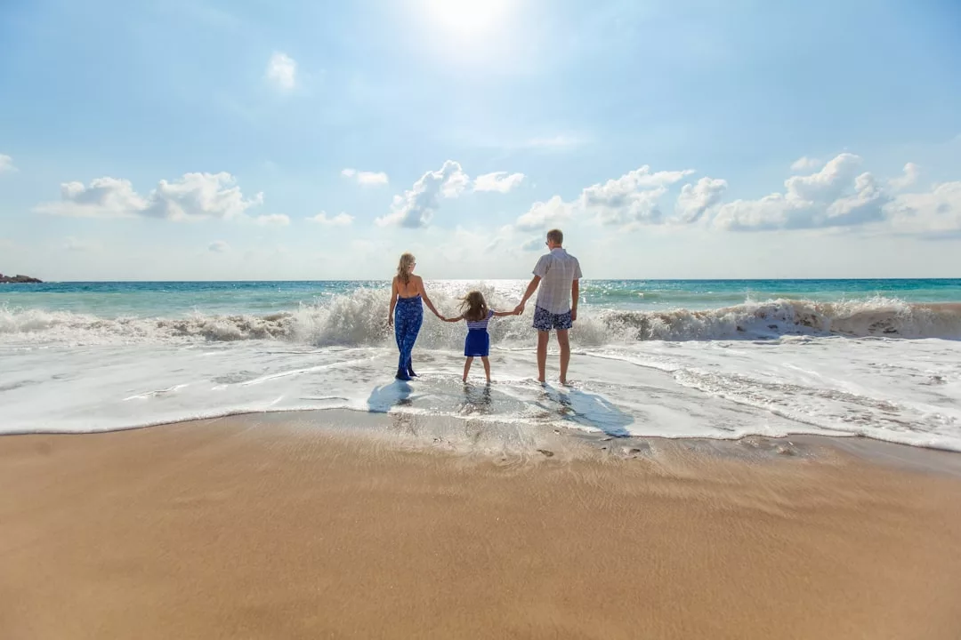 Drei Personen, zwei Erwachsene und ein Kind, stehen händchenhaltend am Rande des Meeres an einem Sandstrand unter einem strahlenden, sonnigen Himmel - ein perfekter Familienurlaub für sonnige Tage und unvergessliche Momente.