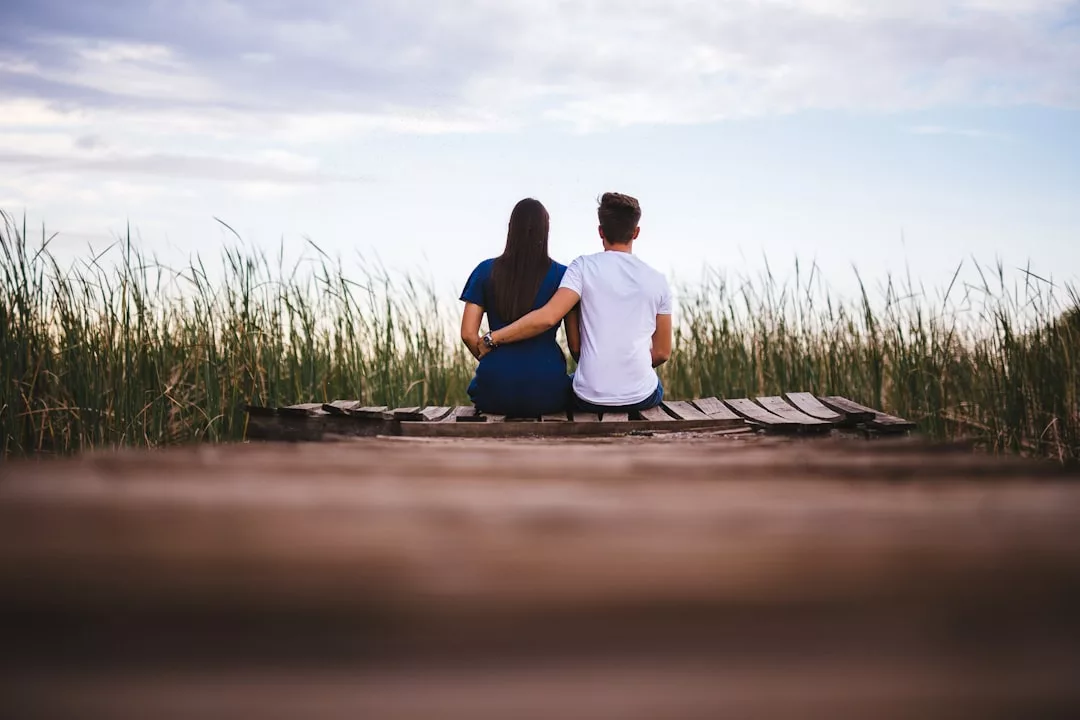 Zwei Menschen sitzen nebeneinander auf einem hölzernen Steg mit Blick auf hohes Gras und einen bedeckten Himmel und teilen einen ruhigen Moment - ein Beweis dafür, dass Ratschläge für eine erfolgreiche Partnerschaft auch in der Nähe wirken.