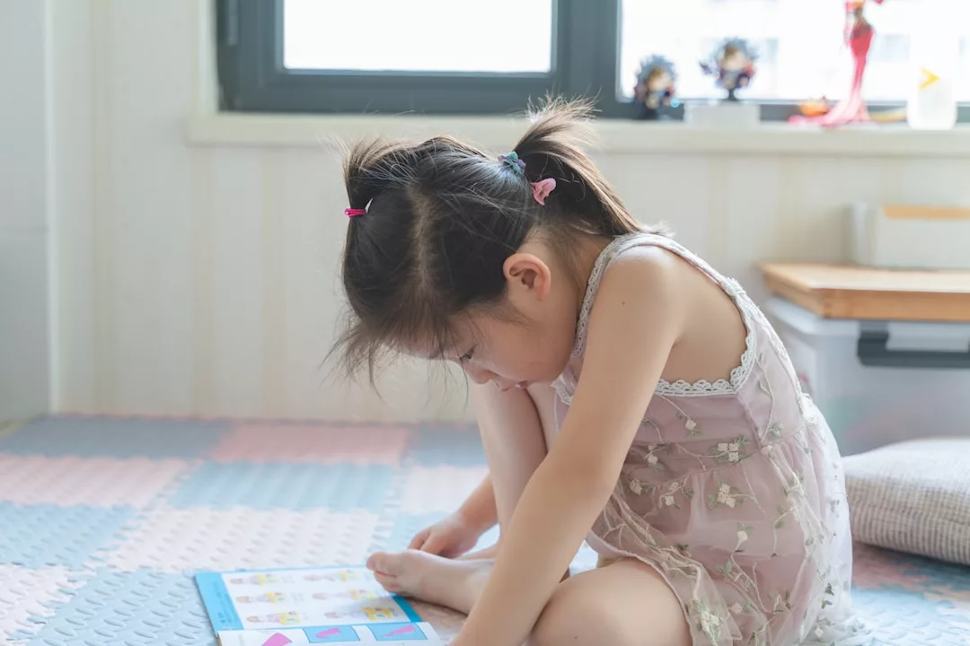 Ein junges Mädchen mit Zöpfen sitzt im Haus auf einer Matte, schaut nach unten und liest in einem Buch, das vor ihr auf dem Boden liegt - ein schönes Beispiel für effektives Homeschooling und Bildung zu Hause.