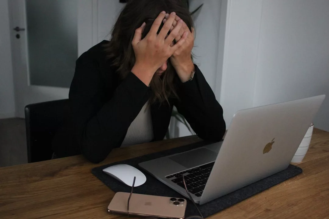 Person sitzt am Schreibtisch mit dem Kopf in den Händen, wirkt gestresst. Auf dem Tisch stehen Smartphone, Brille und eine Tasse - Zeit für Tipps zur Work-Life-Balance, um wieder ausgeglichen durch den Tag zu kommen.
