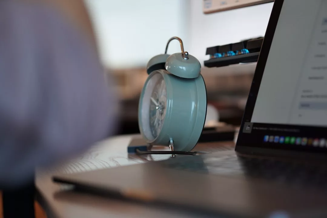 Eine Nahaufnahme eines blauen analogen Weckers auf einem Schreibtisch neben einem Laptop, der Tipps für eine ausgewogene Work-Life-Balance symbolisiert, mit verschwommenen Objekten im Hintergrund.