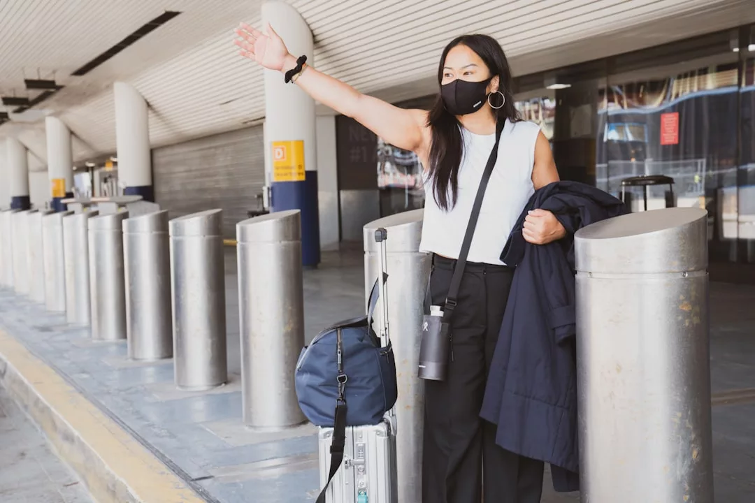Eine Frau mit Gesichtsmaske steht mit Gepäck vor der Tür und hebt den Arm, als wolle sie eine Mitfahrgelegenheit signalisieren. Sie zeigt sicheres Alleinreisen für Frauen und praktische Reisetipps in jeder Situation.