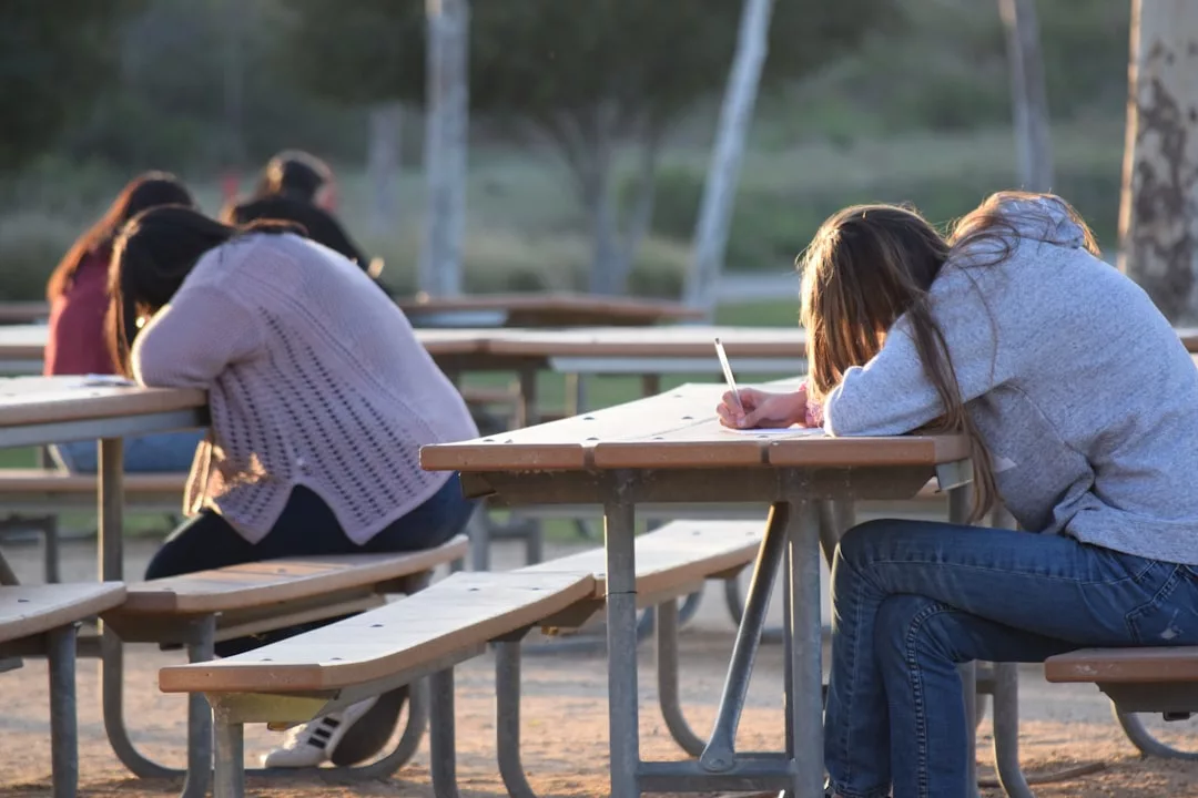 Mehrere Personen sitzen an Picknicktischen im Freien, einige mit gesenktem Kopf und eine Person schreibt auf einem Papier, in einer parkähnlichen Umgebung - vielleicht diskutieren sie gemeinsam Tipps gegen Schulstress oder Wege zur Bewältigung von Schulstress.