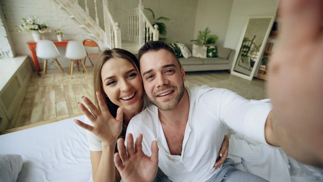Ein Mann und eine Frau sitzen auf einem Bett, lächeln und winken in die Kamera in einem hellen, modernen Wohnraum und geben Tipps zu Fernbeziehungen und wie man sie meistern kann.
