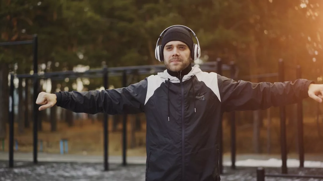 Mann in schwarzer Jacke und mit Kopfhörern streckt seine Arme in einem Outdoor-Fitnessbereich mit Bäumen im Hintergrund waagerecht aus, konzentriert auf seine Fitnessziele und inspiriert von praktischen Fitness-Tipps.