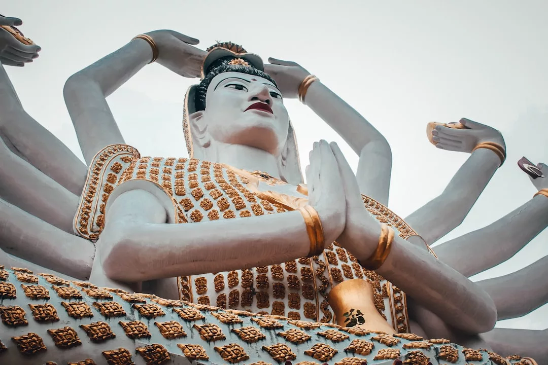 Große Statue einer mehrarmigen Gottheit, deren Hände Wege, Ziele zu setzen und Lebensziele zu erreichen symbolisieren, verziert mit goldenen Mustern und fotografiert aus einem niedrigen Winkel gegen einen hellen Himmel.