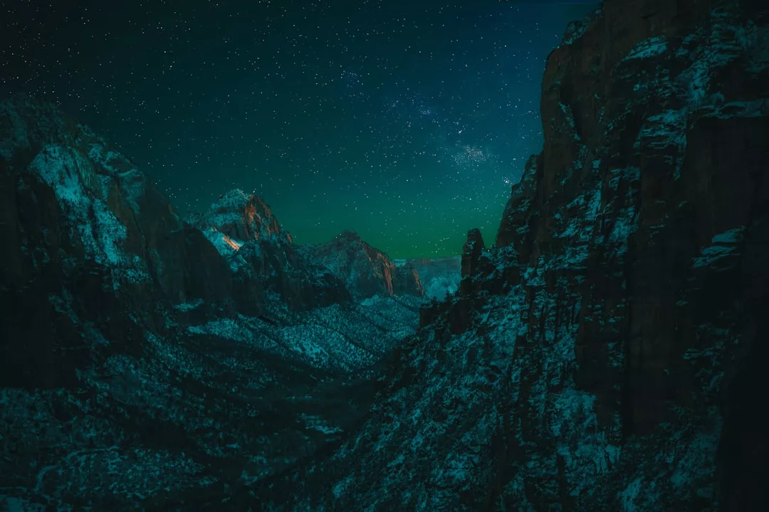 Ein verschneites Gebirgstal unter einem sternenklaren Nachthimmel, mit schroffen Gipfeln und einem schwachen grünlichen Schimmer am Horizont - ein Traumbild für Landschaftsfotografie und alle, die Tipps für perfekte Bilder in der Natur suchen.