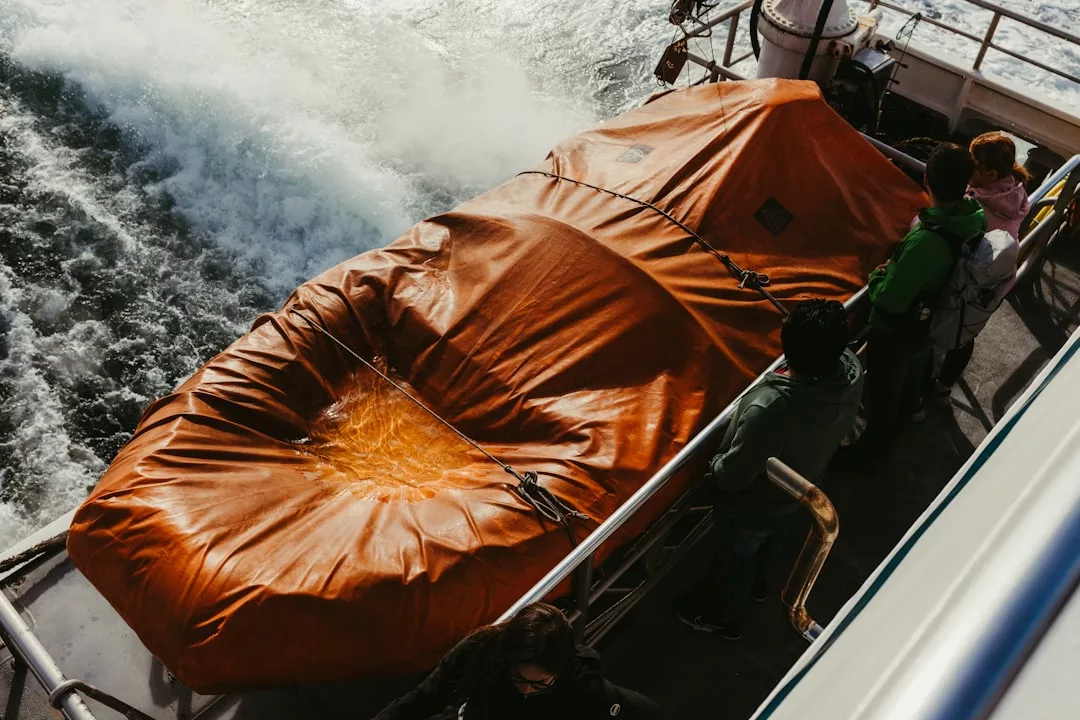 Eine Gruppe von Personen, die sich mit der Reisesicherheit beschäftigen, steht in der Nähe eines orangefarbenen, mit einer Plane abgedeckten Objekts auf dem Deck eines fahrenden Bootes, wobei das Wasser im Hintergrund zu sehen ist.