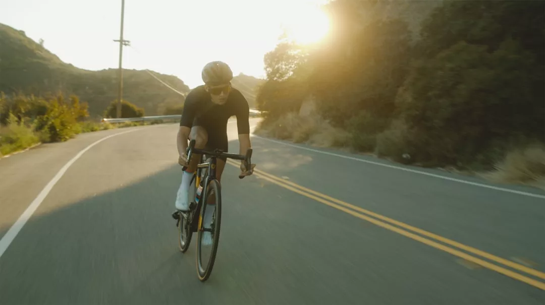 Ein Radfahrer mit Helm fährt mit seinem Rennrad auf einer kurvenreichen Straße bei Sonnenaufgang oder Sonnenuntergang, umgeben von Hügeln und Bäumen - perfekt für alle, die ihre Ausdauer steigern und ihr Durchhaltevermögen mit effektiven Methoden verbessern wollen.