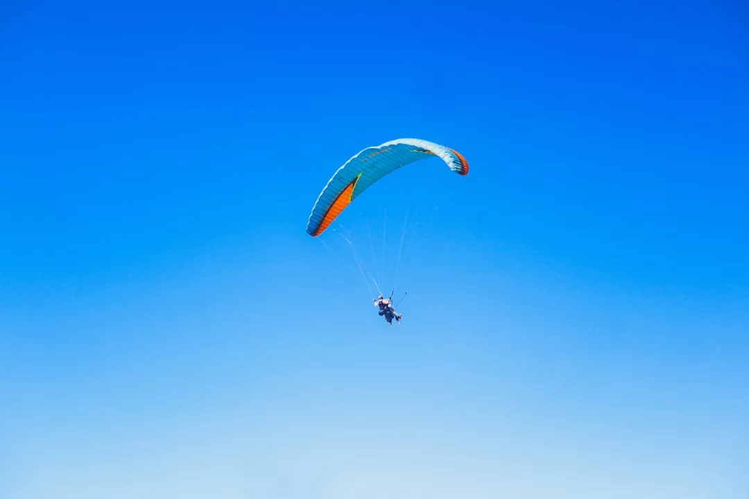 Eine Person genießt den Abenteuersport Gleitschirmfliegen unter einem blauen und orangefarbenen Fallschirm vor einem klaren blauen Himmel - perfekt für alle, die Top Reiseziele suchen.