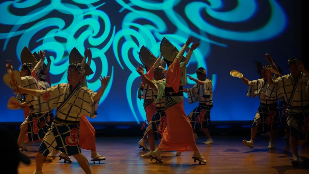 Eine Gruppe von Tänzern in traditionellen japanischen Kostümen tritt auf der Bühne auf, während im Hintergrund blaue, gemusterte Beleuchtung Traditionelle Tänze und Tänze aus aller Welt zeigt.