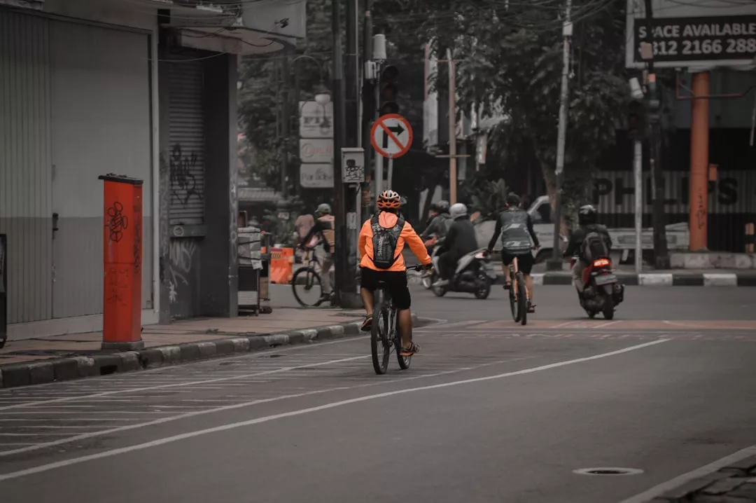 Ein Radfahrer in einer orangefarbenen Jacke fährt eine Straße in der Stadt entlang, gefolgt von anderen und einem Motorrad an einem bewölkten Tag - perfekt für kreative Inspirationen oder die Entdeckung neuer Upcycling-Ideen inmitten der urbanen Energie.