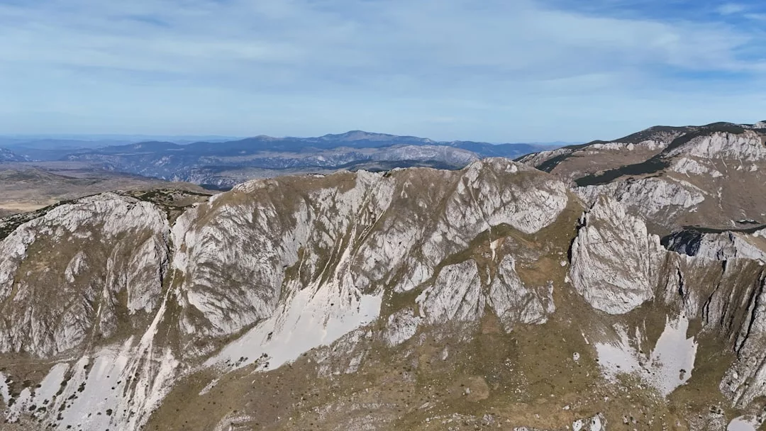 Eine zerklüftete Bergkette mit felsigen, unebenen Hängen und spärlicher Vegetation unter einem teilweise bewölkten Himmel - perfekt für Urlaub in den Bergen. Entfernte Hügel und Berge erstrecken sich über den Horizont für eine friedliche Auszeit in der Natur.