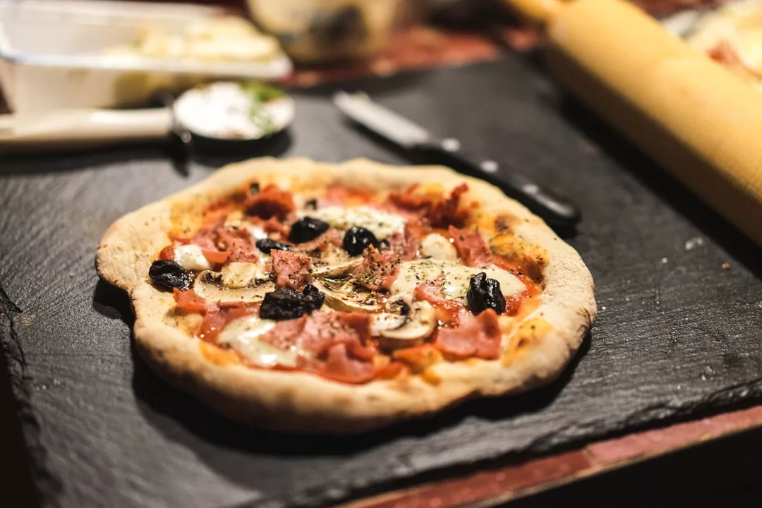 Eine frisch gebackene Pizza mit Pilzen, schwarzen Oliven und Schinken liegt auf einer Schiefertafel - perfekt zum Teilen, während Sie Beziehungskommunikation und Kommunikation verbessern. Im Hintergrund liegen ein Messer und ein Nudelholz.