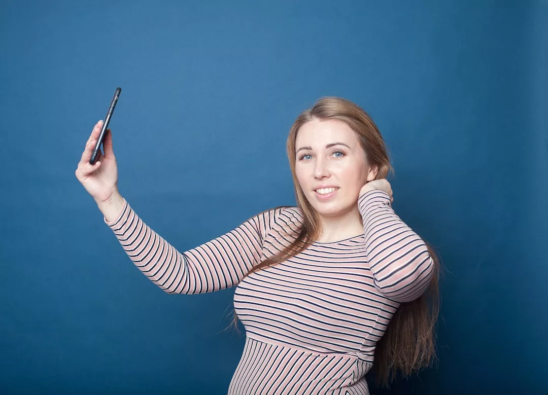 Eine Frau mit langen Haaren und gestreiftem Hemd macht ein Selfie mit einem Smartphone und hebt dabei moderne Kommunikation vor einem schlichten blauen Hintergrund hervor.