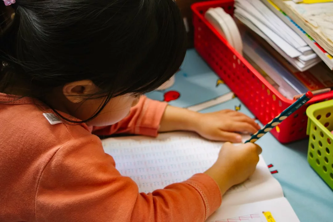 Ein Kind in einem orangefarbenen Hemd arbeitet an Lernfähigkeiten, während es an einem Schreibtisch mit Körben, die Papiere und Bücher enthalten, in ein Arbeitsbuch schreibt.