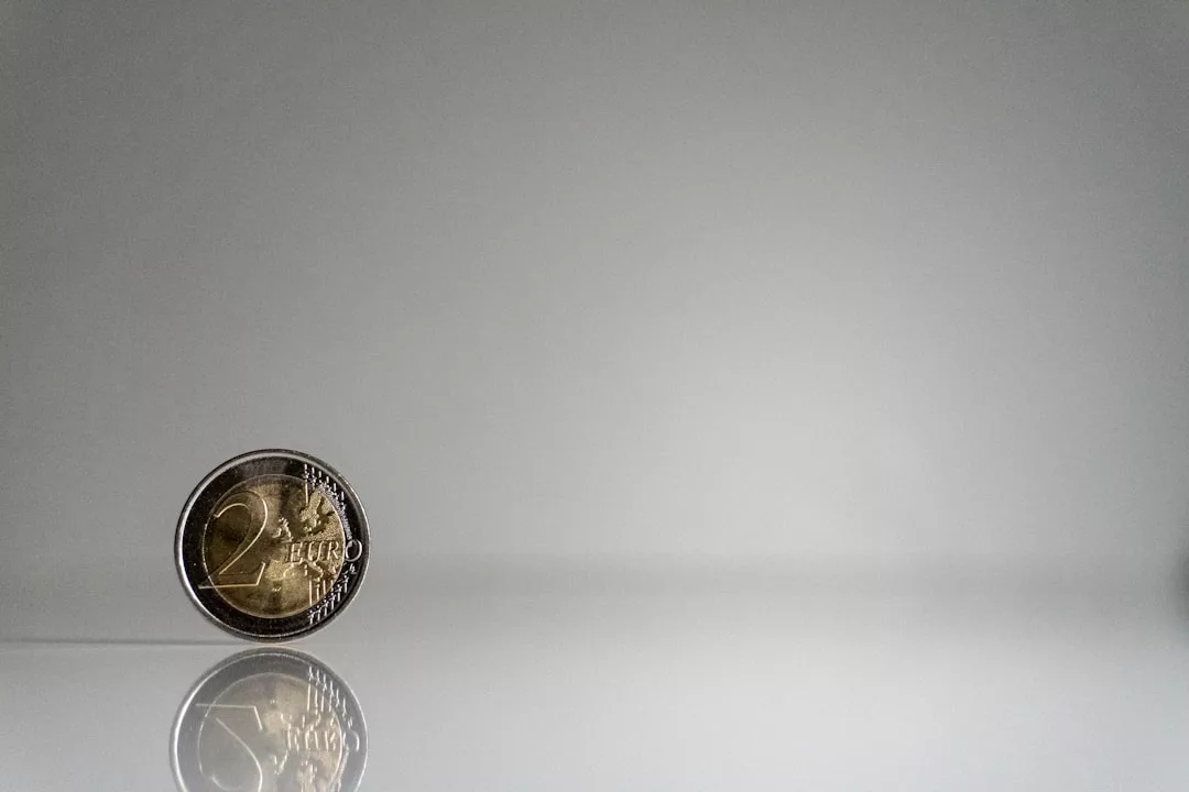 Eine aufrecht stehende 2-Euro-Münze auf einer spiegelnden weißen Oberfläche mit einem schlichten grauen Hintergrund, die Vermögensaufbau und Strategien für wirtschaftlich unsichere Zeiten symbolisiert.