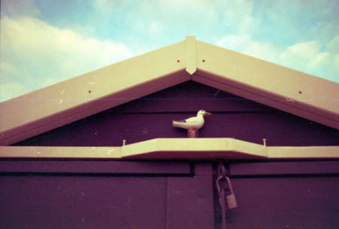 Eine kleine weiße Vogelfigur hockt auf einem Regal unter der Dachspitze einer dunklen Holzkonstruktion, darunter ein Vorhängeschloss und darüber ein teilweise bewölkter Himmel - die perfekte Inspiration für alle, die Vogelhäuser bauen und nach Tipps oder Anleitungen suchen.