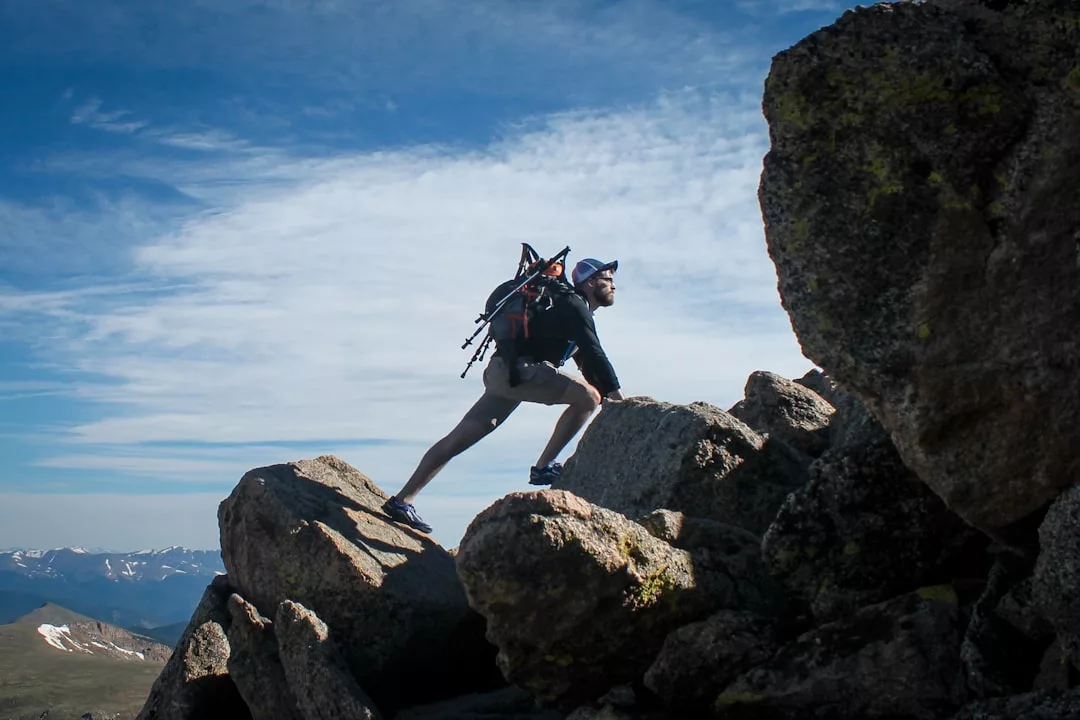 Eine Person mit Rucksack und Mütze wandert über große, felsige Brocken eines Berges, genießt dabei die Natur und erblickt ferne Gipfel unter einem teils bewölkten Himmel.