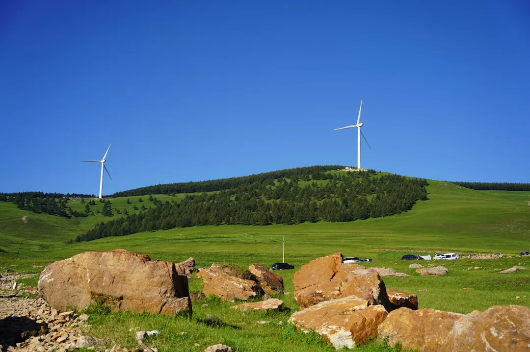 Zwei Windkraftanlagen, Symbole für Effizienz und Zuverlässigkeit erneuerbarer Energieanlagen, stehen auf einem grasbewachsenen, baumbestandenen Hügel unter klarem Himmel; im Vordergrund sind große Felsen und parkende Autos zu sehen.