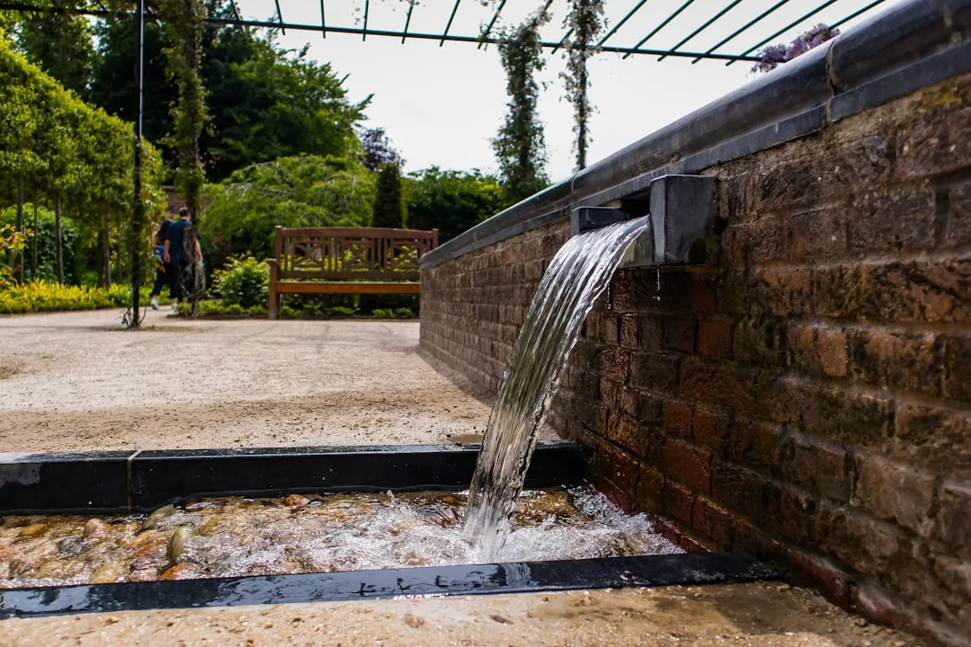 Ein Wasserstrahl fließt aus einem Ausguss in einer Backsteinmauer in eine seichte Rinne im Garten. Eine üppige Bepflanzung und eine Holzbank verleihen dieser entspannenden Außenanlage ihren Charme.