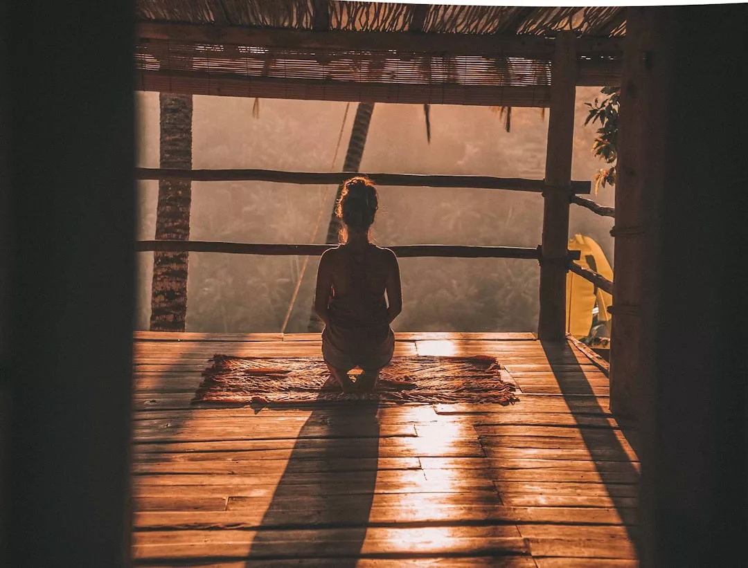 Eine Person sitzt im Schneidersitz auf einer Matte in einer hölzernen Freiluftstruktur bei Sonnenuntergang und genießt die Entspannung, während das Sonnenlicht lange Schatten auf den Boden wirft - eine ruhige Szene, die perfekt für Wellnessreisen und Erholung ist.