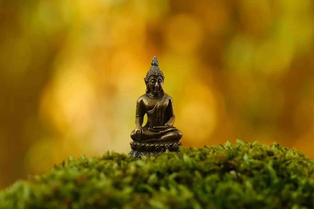 Eine kleine bronzene Buddha-Statue sitzt auf einer moosbewachsenen Fläche mit einem verschwommenen goldgelben Hintergrund und symbolisiert ethische Werte und die Bedeutung von Wertevermittlung in stiller Meditation.