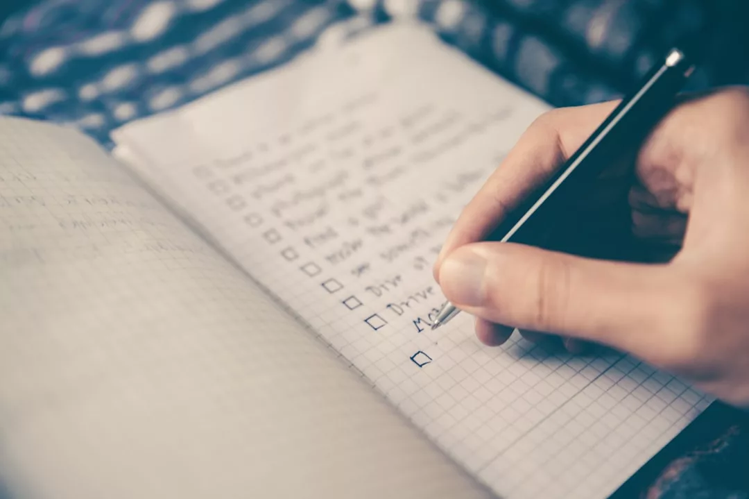 Eine Hand, die einen Stift hält, schreibt auf ein Notizbuch mit einer abgehakten Liste, die mehrere Punkte in einem Gittermuster enthält - perfekt für alle, die ihr Haushaltsbudget planen oder ein effektives Haushaltsbudget erstellen möchten.