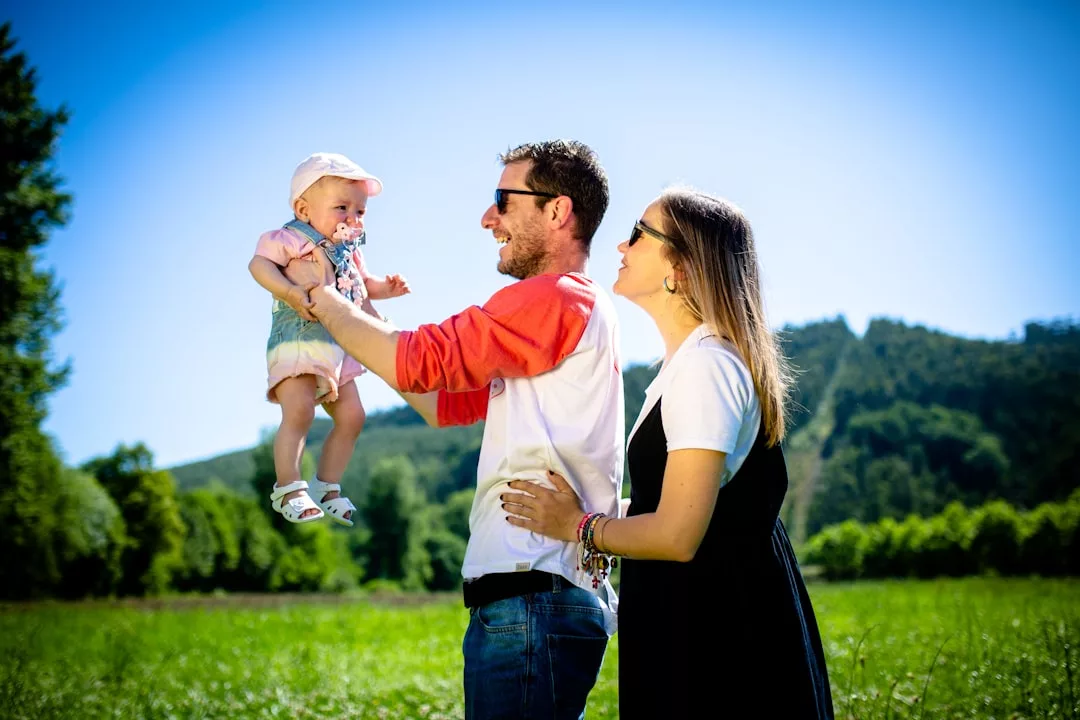 Ein Mann hält ein Baby in die Luft, während er neben einer Frau steht und das Familienleben in einer sonnigen, grasbewachsenen Umgebung mit Bäumen und Hügeln im Hintergrund genießt.