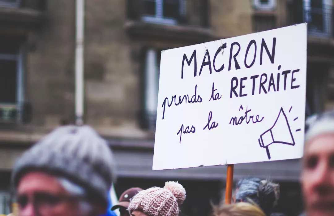 Eine Gruppe von Menschen hält im Freien ein Schild mit der Aufschrift "Macron prends ta retraite pas la nôtre" ("Macron, nimm deinen Ruhestand, nicht unseren") in französischer Sprache, um auf die Bedenken hinsichtlich Ruhestand und finanzieller Vorsorge hinzuweisen.
