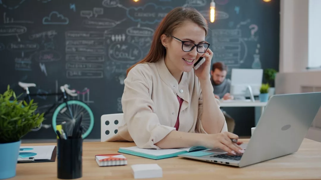 Eine Frau sitzt an einem Schreibtisch, benutzt einen Laptop und telefoniert in einem modernen Büro mit einer Kreidetafelwand und einem Fahrrad im Hintergrund, konzentriert darauf, ihre Produktivität zu steigern und effizient zu arbeiten.