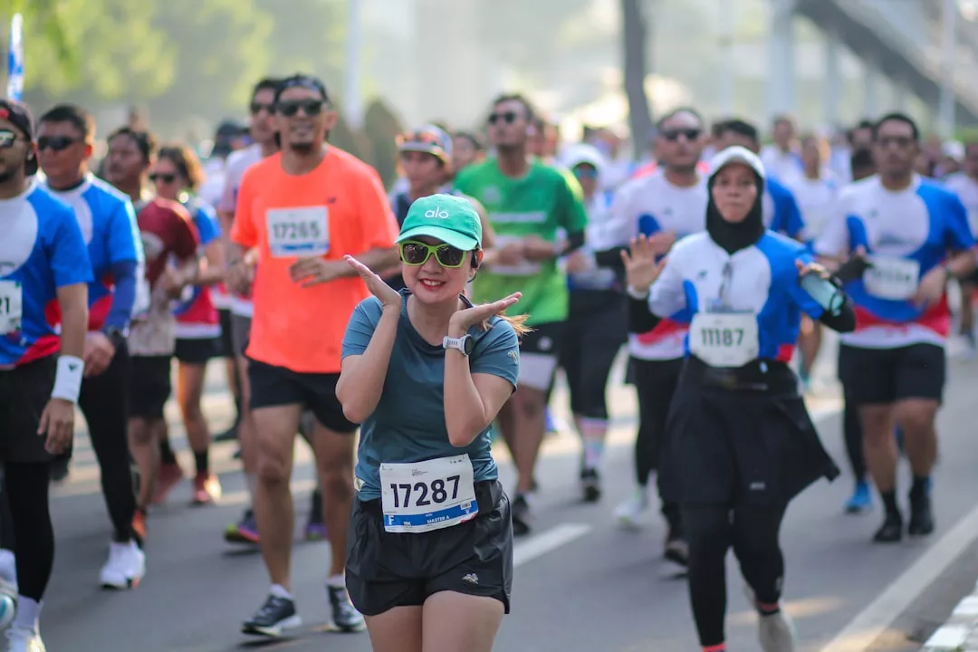 Eine Gruppe von Marathonläufern auf einer Straße in der Stadt; eine Frau mit grüner Mütze und Sonnenbrille posiert mit erhobenen Händen und lächelt in die Kamera - ein Zeichen für die Freude am engagierten Marathontraining.