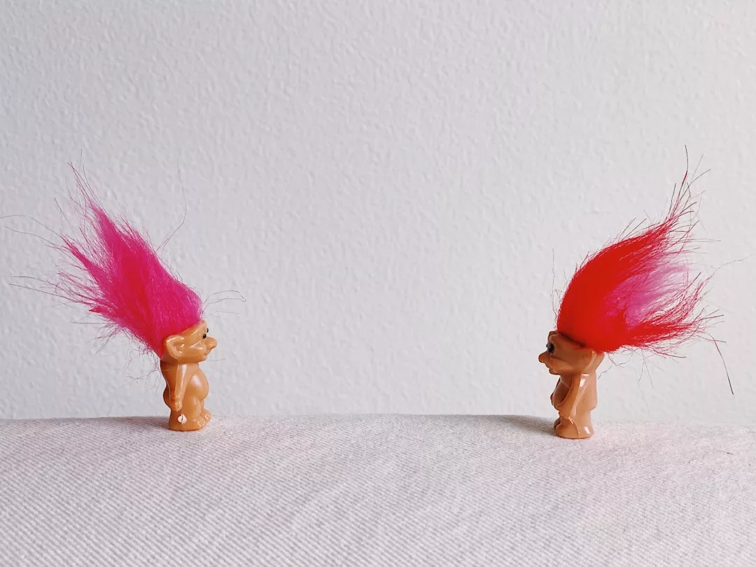 Zwei kleine Trollpuppen mit hellem, aufrechtem Haar - eine rosa und eine rote - stehen sich vor einem schlichten weißen Hintergrund gegenüber und symbolisieren die Wiederannäherung von entfremdeten Familienmitgliedern.