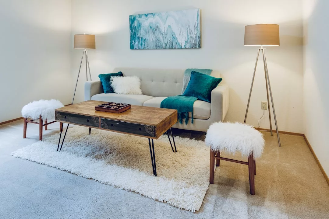 Modernes Wohnzimmer mit einem beigen Sofa, tealfarbenen Akzenten, einem Couchtisch aus Holz, zwei pelzigen Hockern, gemütlichen Wohnaccessoires als Ergänzung, zwei Stehlampen, einem weißen Teppich und abstrakter Wandkunst über dem Sofa. Perfekt für ein stilvolles Zuhause.