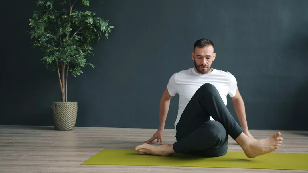 Ein Mann übt einfache Übungen auf einer grünen Yogamatte im Haus, schlägt ein Bein über das andere, während er sich auf einem Holzboden dehnt, mit einer Topfpflanze im Hintergrund - ideal für Anfänger und Yoga-Routinen.