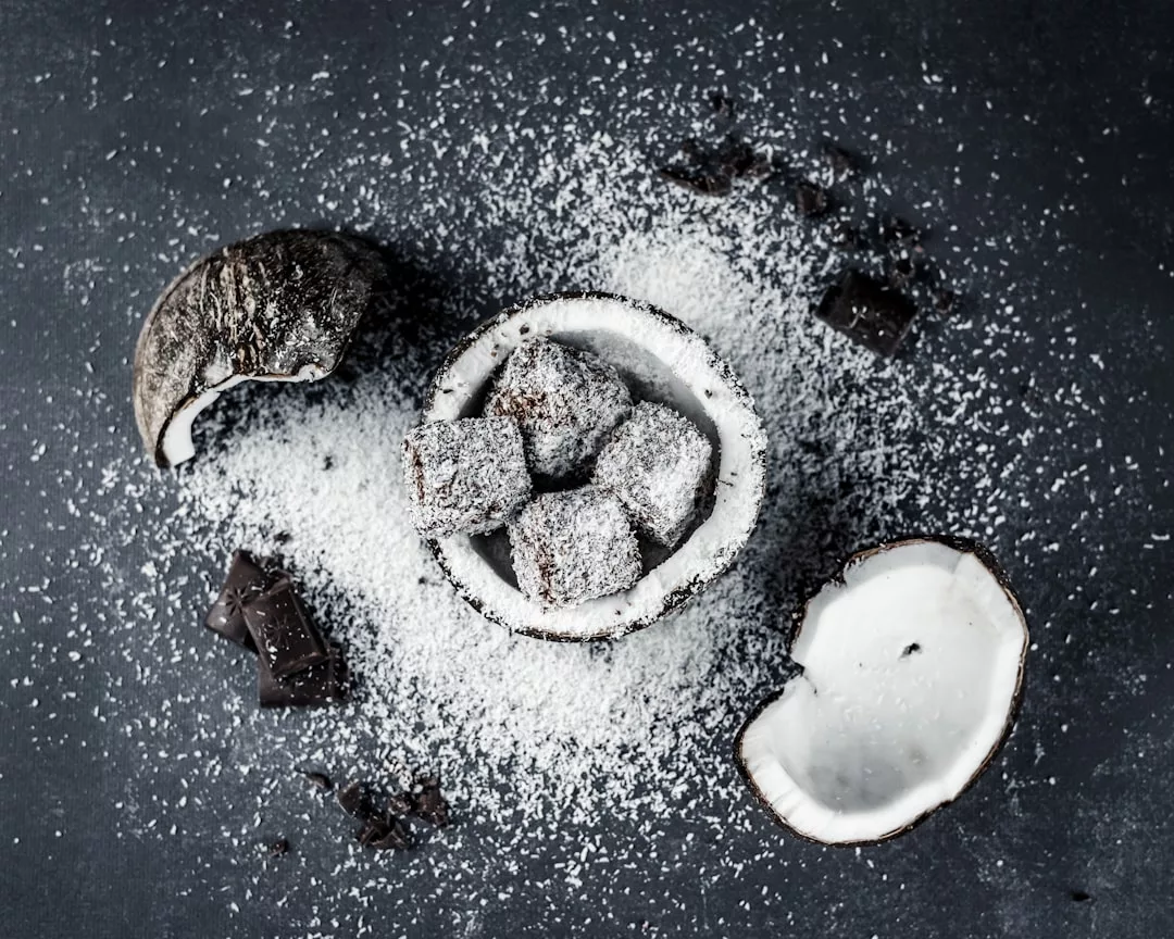 Kokosnussschale gefüllt mit Schokoladenwürfeln, umhüllt von Kokosraspeln, umgeben von Kokosflocken, Schokoladenstücken und Kokosnussschalen auf einer dunklen Oberfläche - perfekt für gesunde Rezepte oder als Inspiration für zuckerfreie Rezepte.